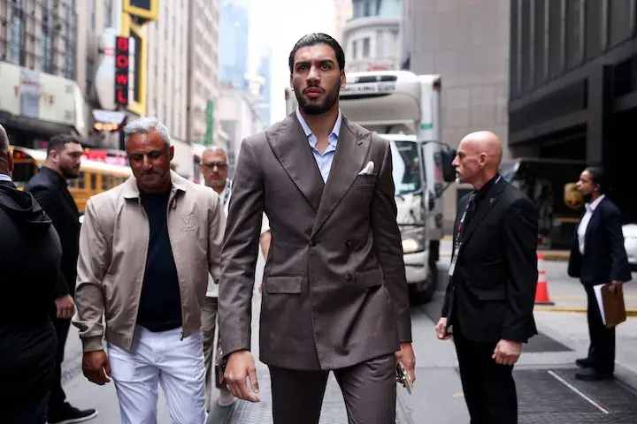 Hamzah Sheeraz is wearing a suit and shown walking through a city street, with multiple people, vehicles and buildings to his side and behind him.
