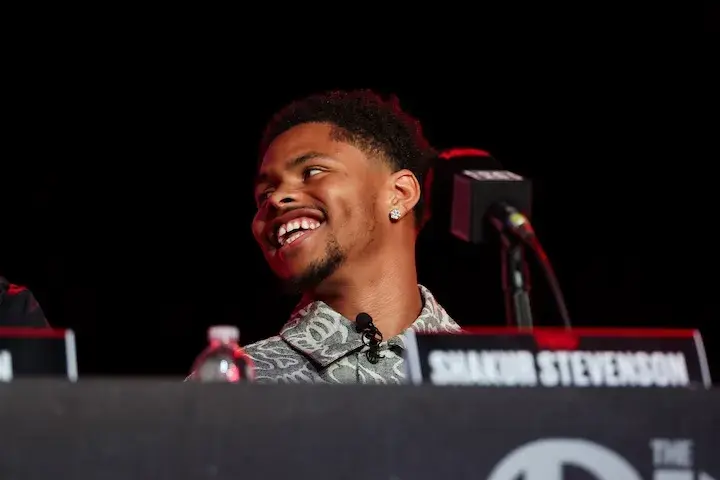 Shakur Stevenson is seated at a podium with his name on a nameplate in front of him. He is looking to his right and smiling.