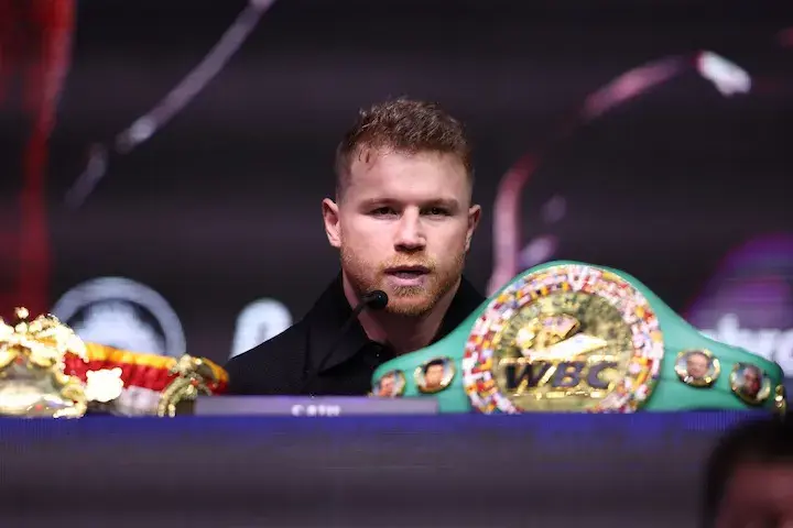 Canelo Alvarez sits in front of a dais with the WBC super middleweight and Ring Magazine super middleweight championship belts in front of him.