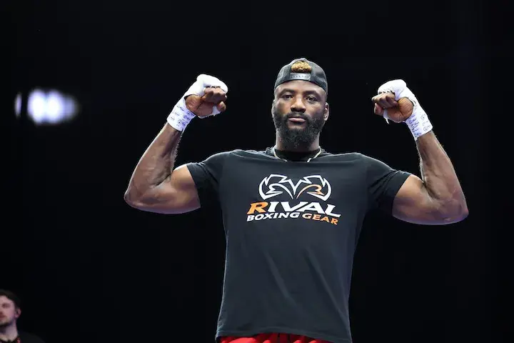 Heavyweight contender Efe Ajagba stands with his hands up and his biceps flexed while wearing a black T-shirt that says Rival Boxing Gear.
