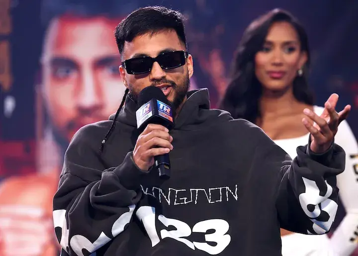 Junior featherweight contender Ramon Cardenas sits while holding a microphone and wearing a black-and-white hooded sweatshirt. A woman in a white dress stands behind him in the background.