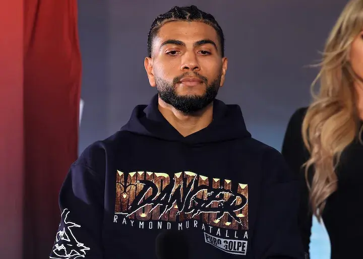 Raymond Muratalla sits in a hooded black sweatshirt that says "Danger" in large letters with "Raymond Muratalla" underneath. A woman with long hair can be partially seen standing slightly behind him to his left.