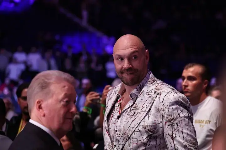 Tyson Fury, wearing a suit, stands in an arena near boxing promoter Frank Warren.